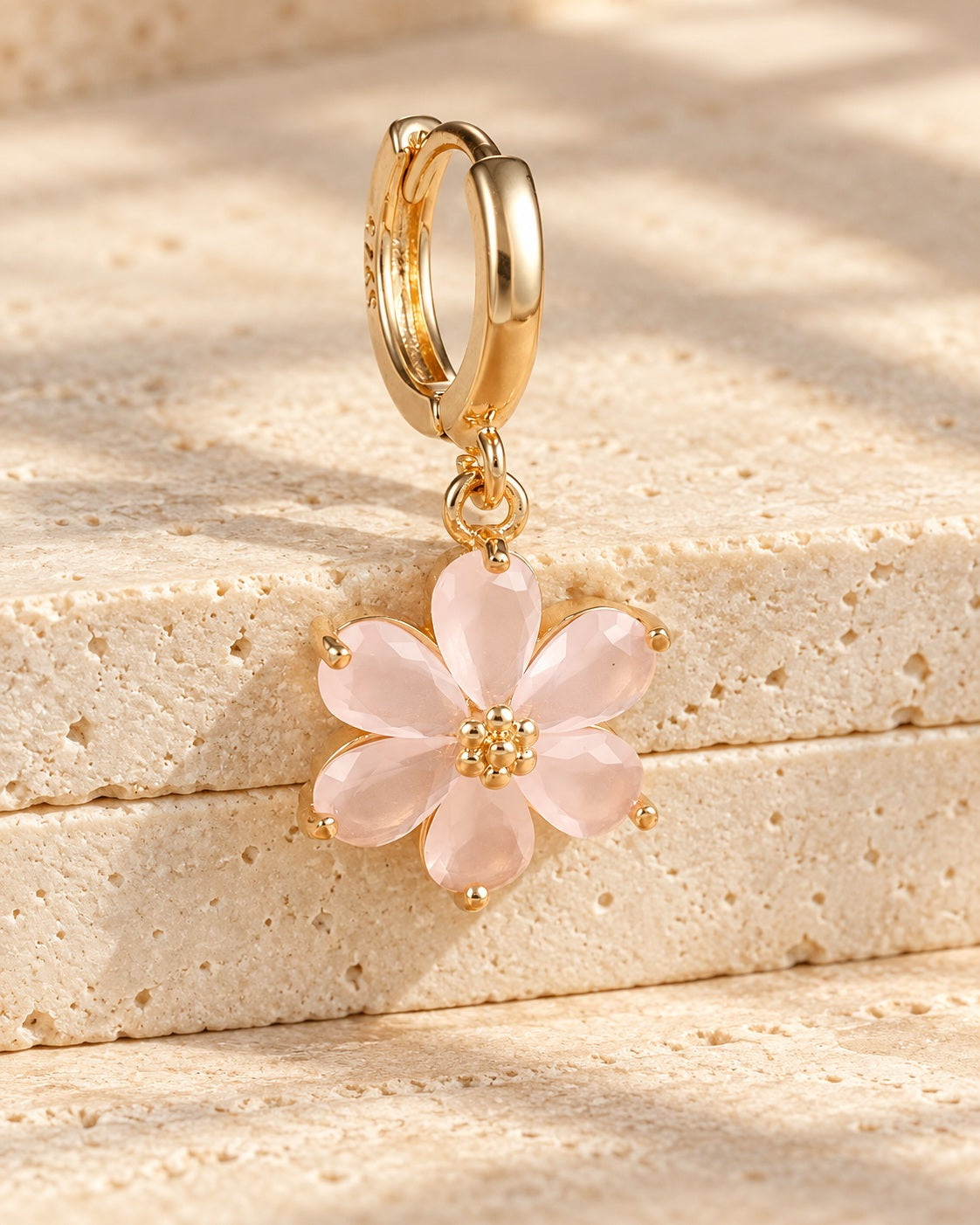 Gold-tone mini hoop with a pale-pink six-petal flower drop on textured warm stone